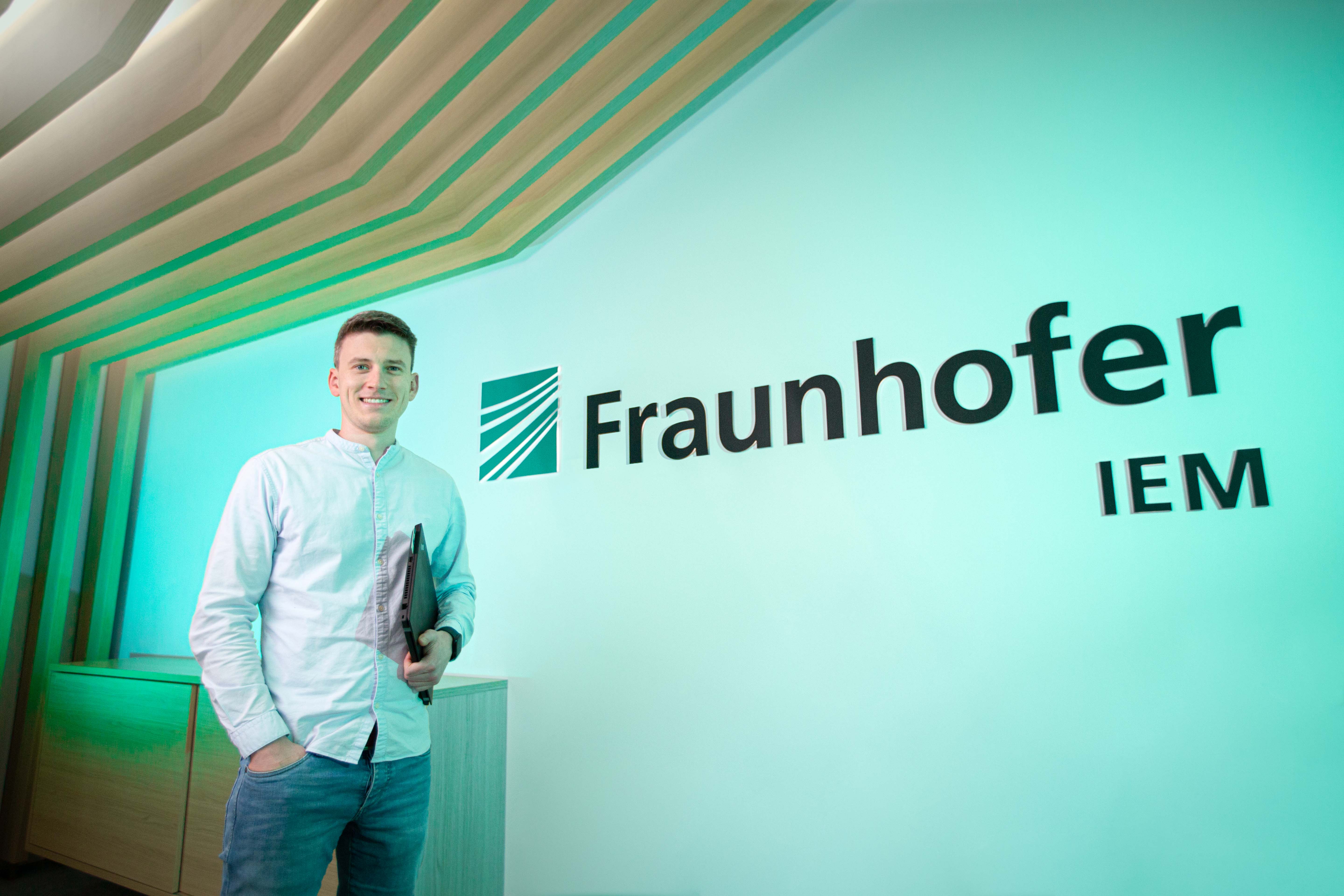 Christian Kürpick steht mit einem Laptop in der Hand vor einem Fraunhofer Logo und Schriftzug.