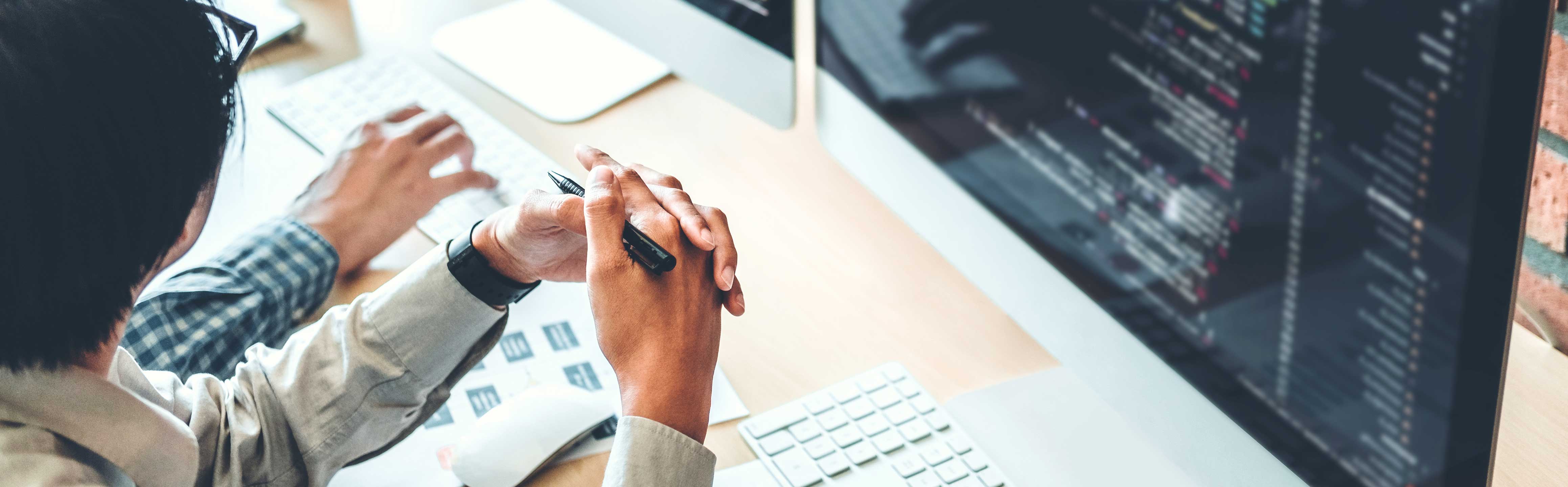 Anschnitt von zwei Personen am Schreibtisch vor einem Computerbildschirm, von der eine Person einen Stift in den Händen hält.