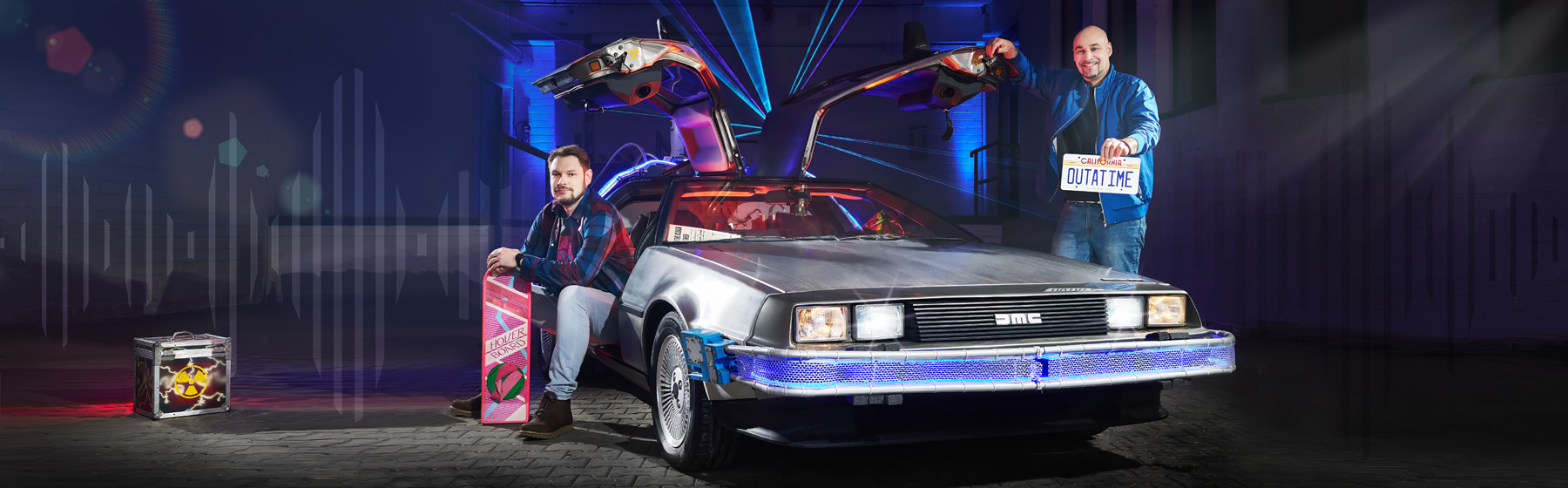 Zwei Männer posieren vor einem DeLorean mit geöffneten Flügeltüren, inspiriert von „Zurück in die Zukunft“. Einer sitzt mit einem Hoverboard, der andere hält ein Nummernschild mit der Aufschrift „OUTATIME“.