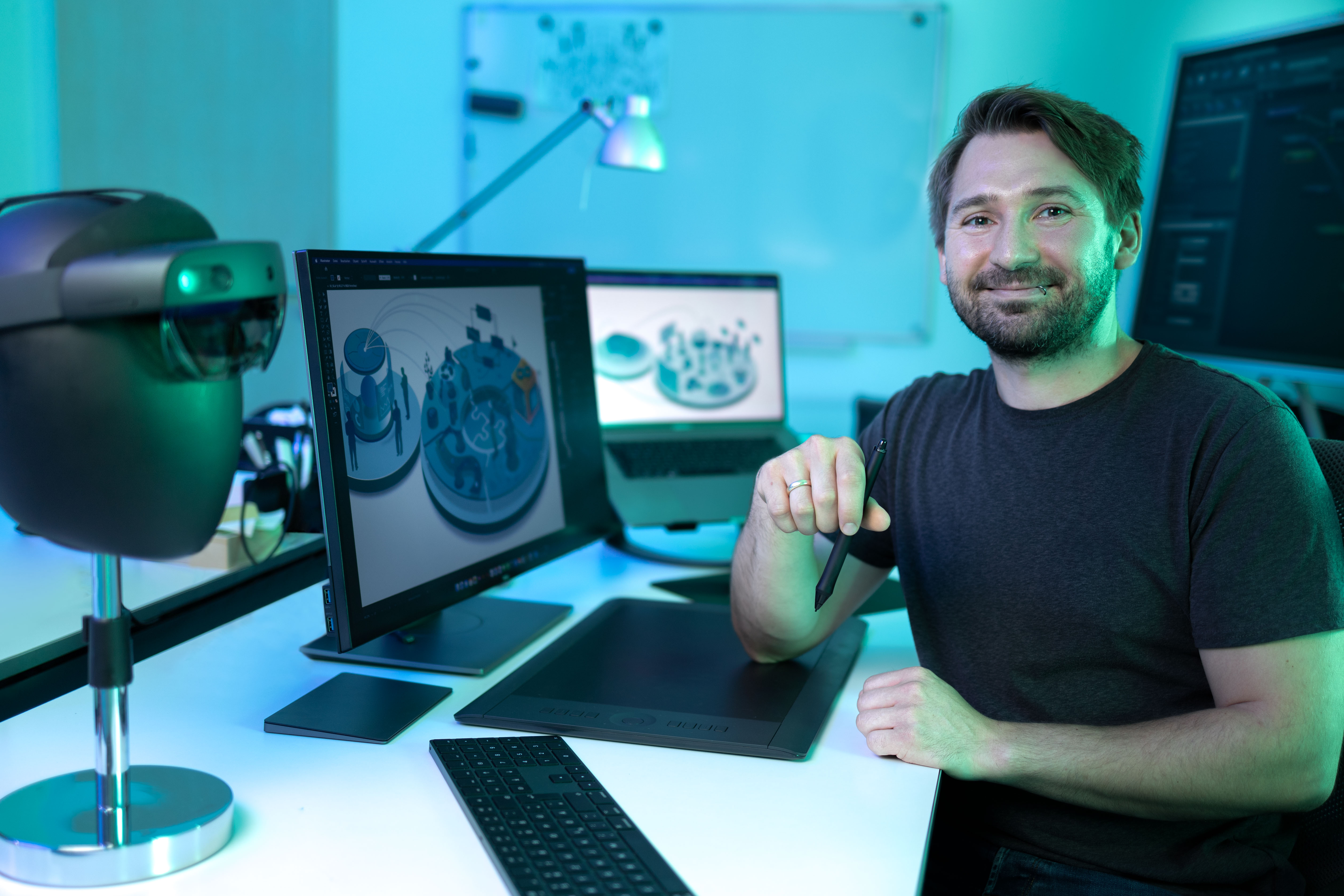 William Neufeld sitzt lächelnd in seinem Büro, während auf seinen Computern Grafikanwendungen im Betrieb sind.