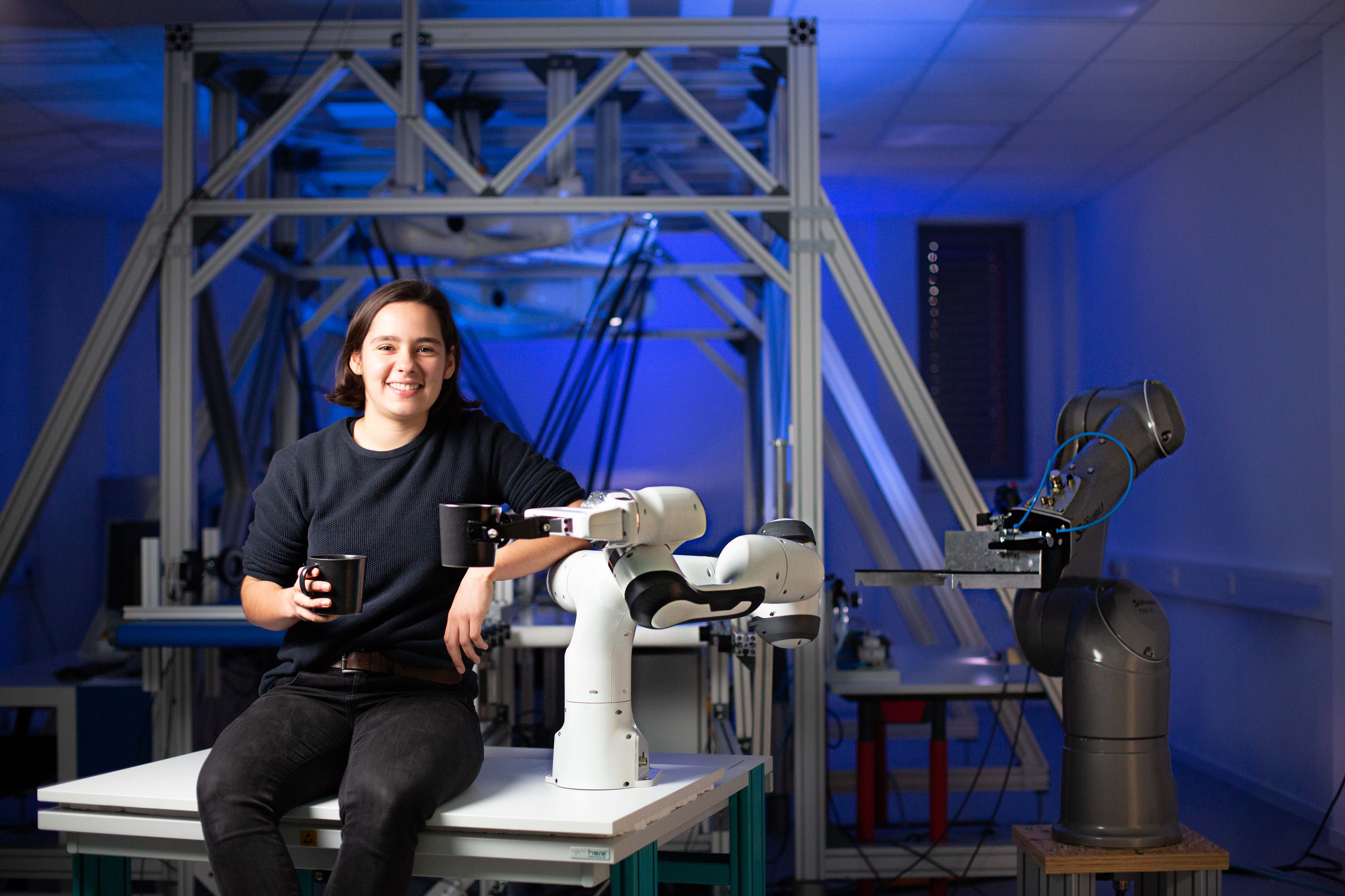 Gerit Saliger sitzt angelehnt an einen Robotergreifarm mit einer Tasse in der Hand. Der Roboterarm hält ebenfalls eine Tasse in der Hand.