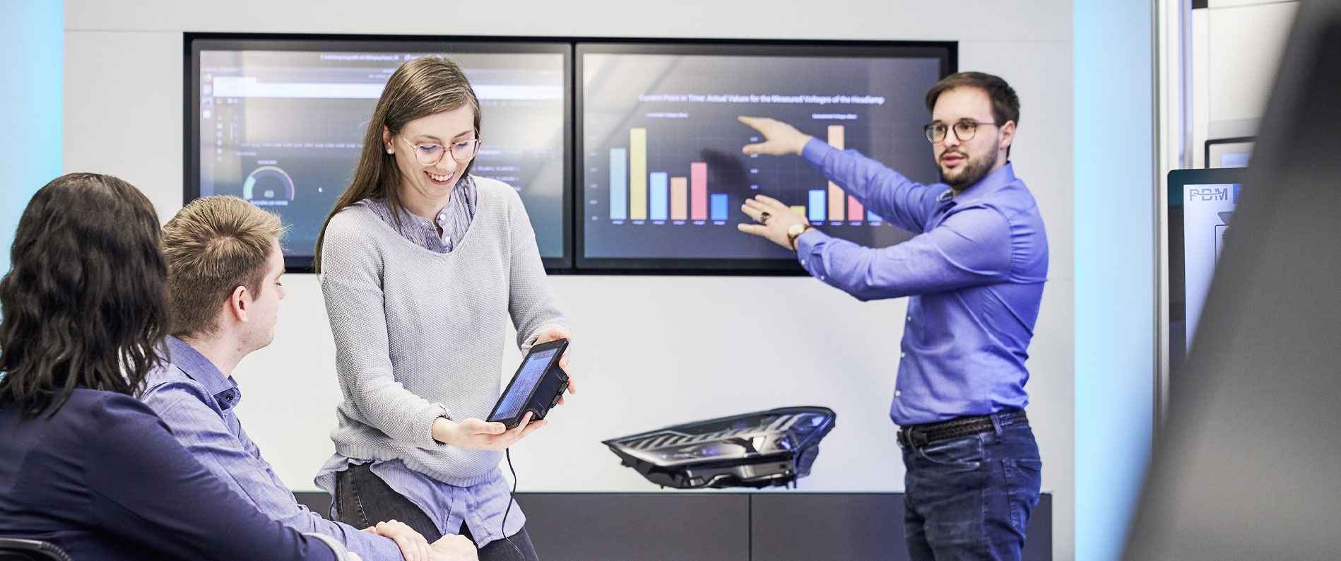 Frau zeigt zwei Personen Daten auf dem Tablet, während Mann auf Säulendiagramm am Bildschirm zeigt