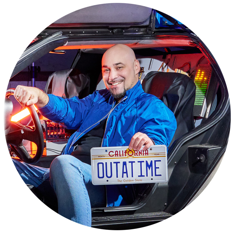 Ein lächelnder Mann in einer blauen Jacke sitzt am Steuer eines DeLorean mit geöffneter Tür. Er hält ein kalifornisches Nummernschild mit der Aufschrift „OUTATIME“ in der Hand. Im Hintergrund sind bunte Lichter und technische Details des Fahrzeugs zu sehen.