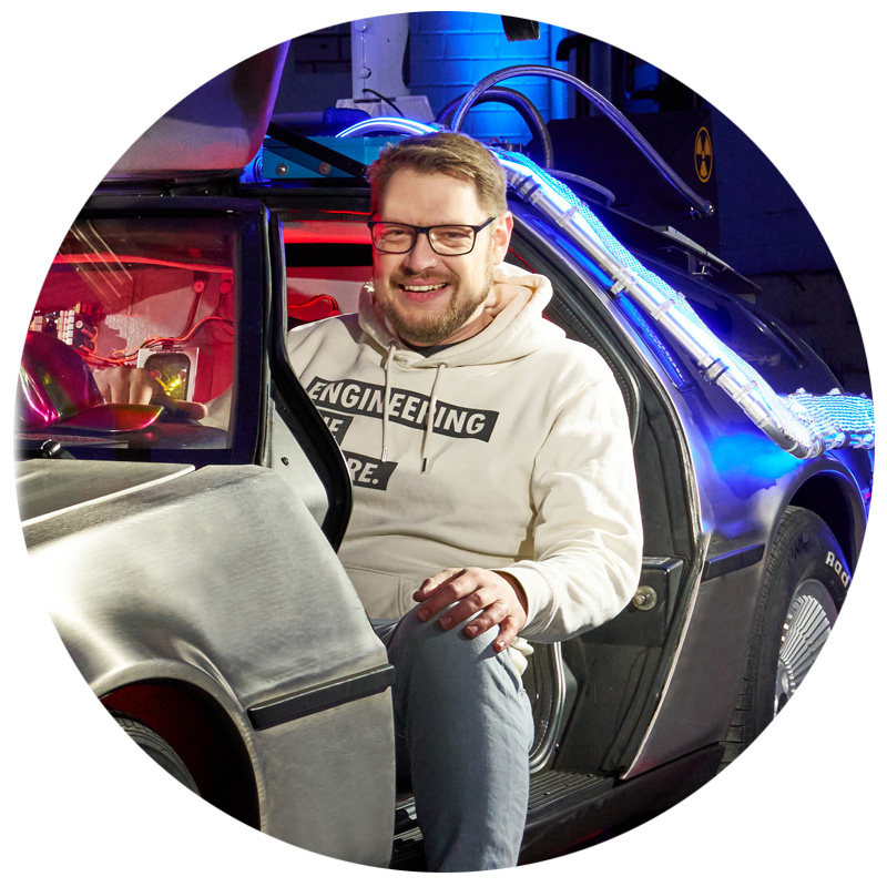Ein lachender Mann mit Brille und hellgrauem Hoodie, auf dem „ENGINEERING THE FUTURE“ steht, sitzt in der offenen Tür eines DeLorean. Im Hintergrund leuchten blaue Lichtstreifen und technische Details des Fahrzeugs sind sichtbar.