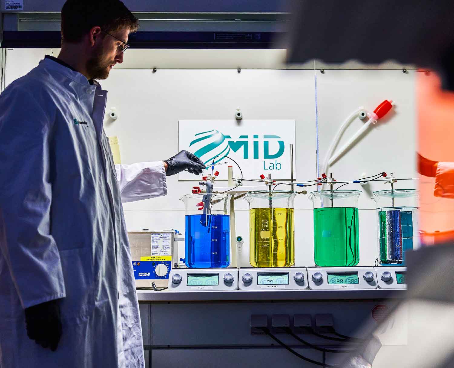 Ein Mann steht in einem Laborkittel vor vier großen Gefäßen mit farbigen Flüssigkeiten und taucht ein Bauteil in ein Gefäß ein