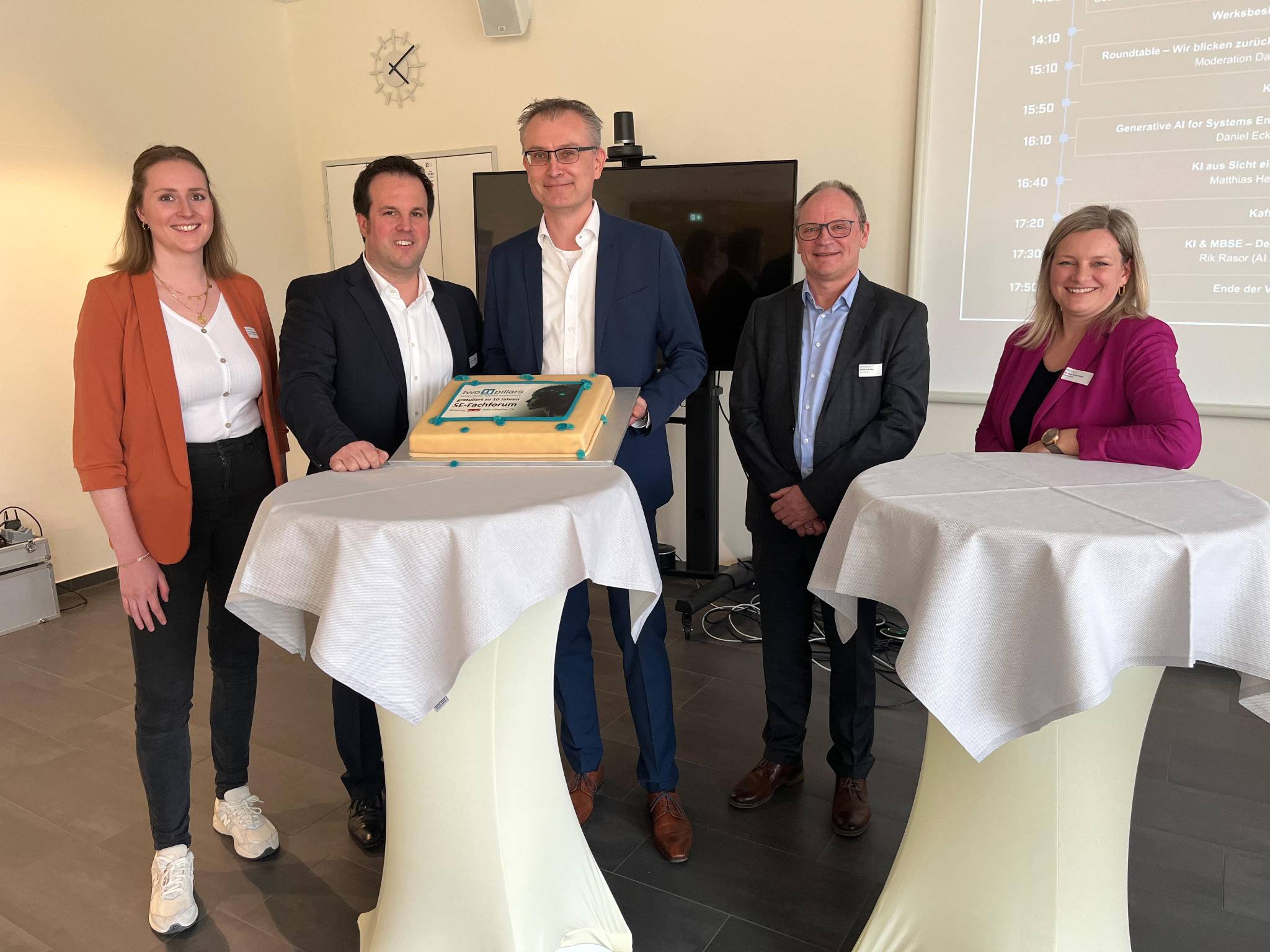 Fünf Menschen stehen an zwei Hochtischen um einen Kuchen herum.