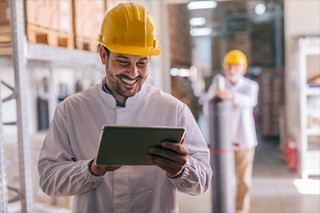 Mann mit Warnweste und Sicherheitshelm in einer Lagerhalle schaut auf ein Tablet.