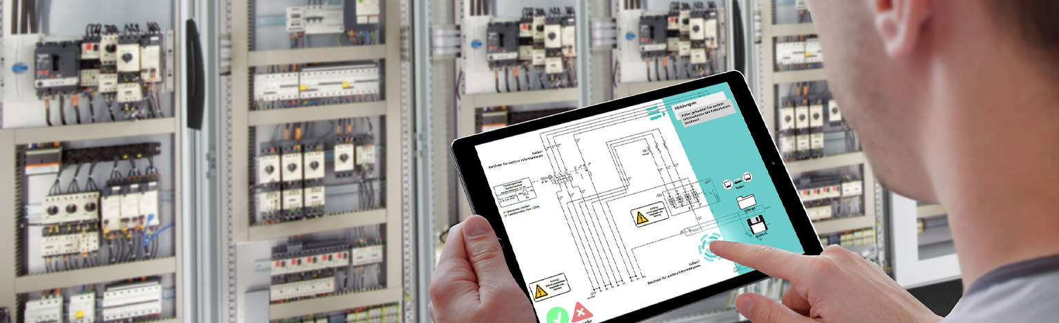 Tablet in einer Industriehalle zeigt den Plan einer Schaltanlage, eine Hand zeigt darauf.