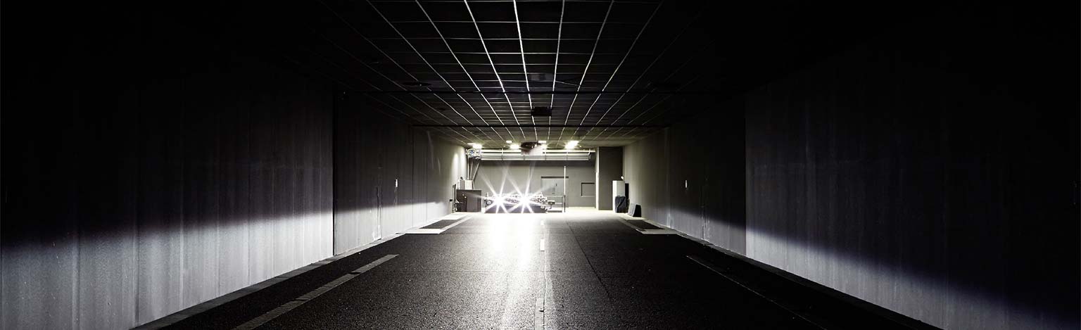 Eine tunnelartige Straße, welche in eine Industriehalle führt, in der helle Scheinwerfer zu sehen sind..