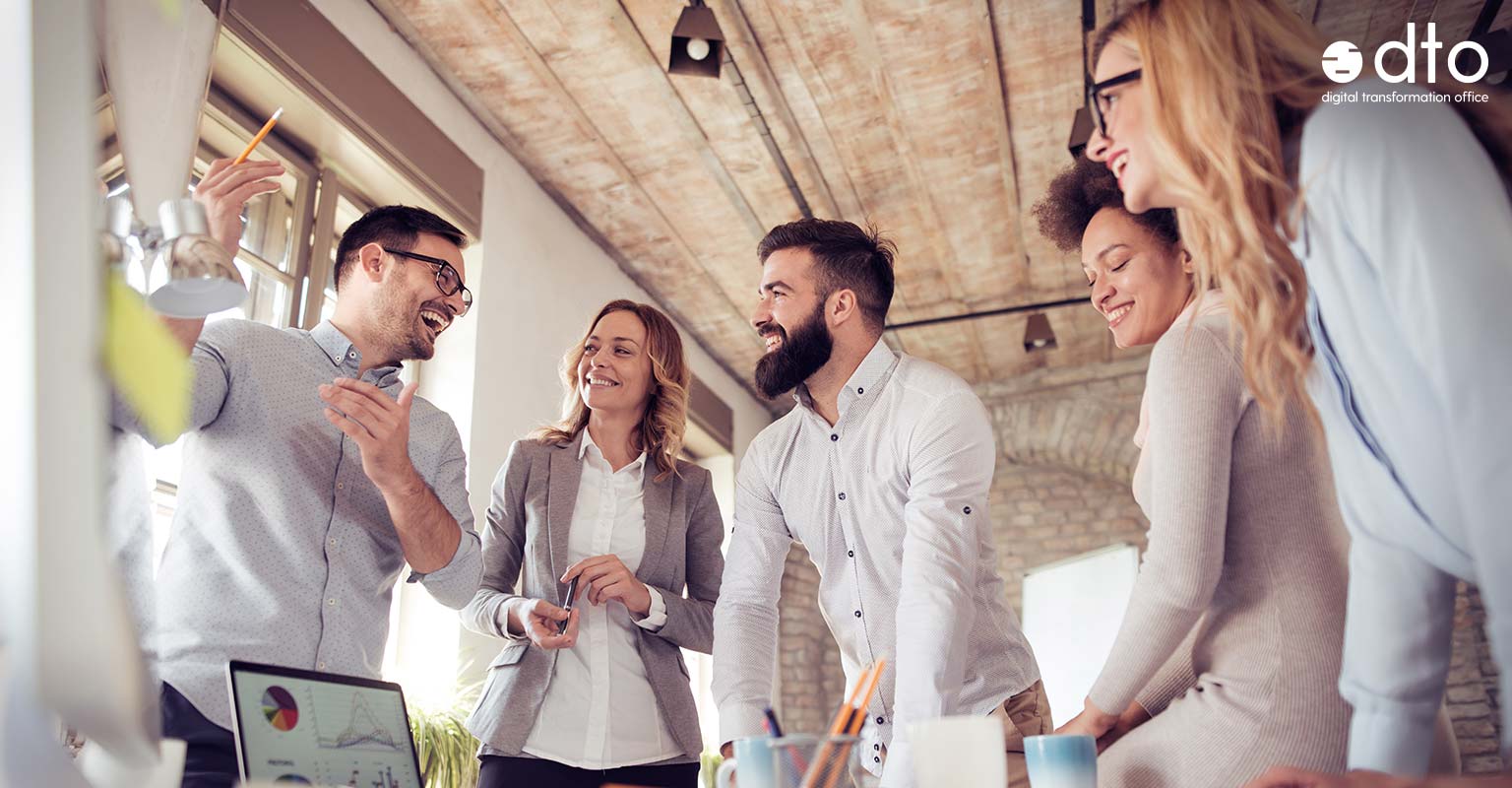 Gruppe von Personen in einem Büro. Eine Person präsentiert etwas, die anderen sind gut gelaunt und lachen.