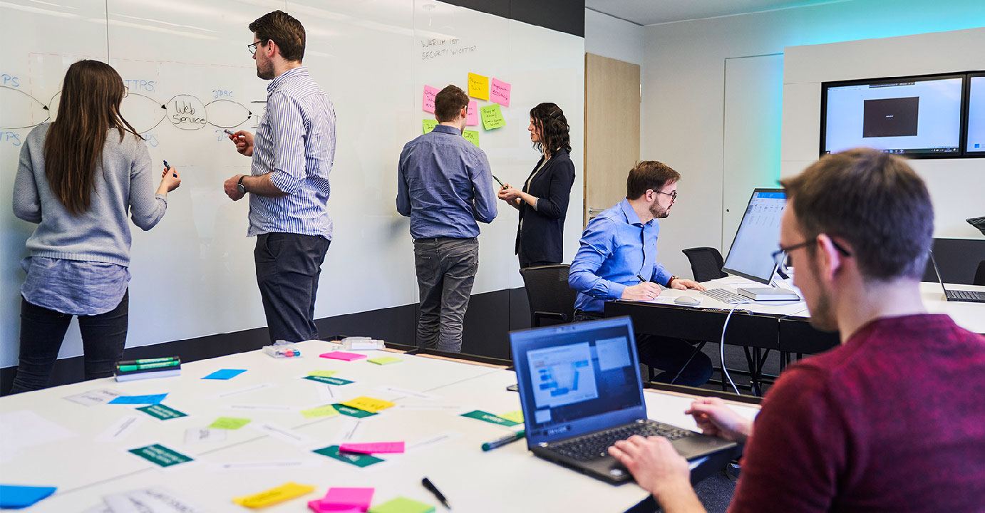 Sechs Personen arbeiten im Secure Engineering Lab verteilt an Tischen und Whiteboards.