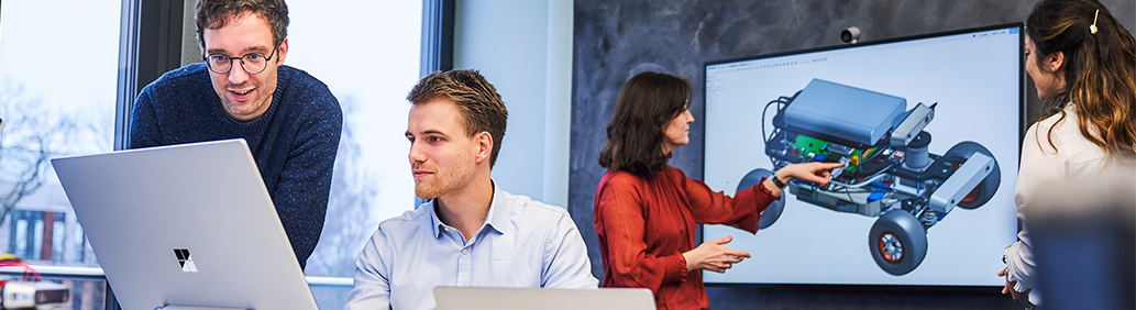 Two men at a computer, behind them two women at a large screen with model illustration.