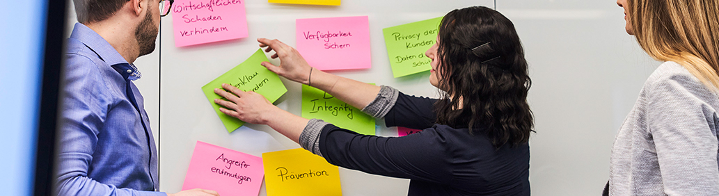 Three people in front of a bulletin board. One of them hangs Post-Its on it.