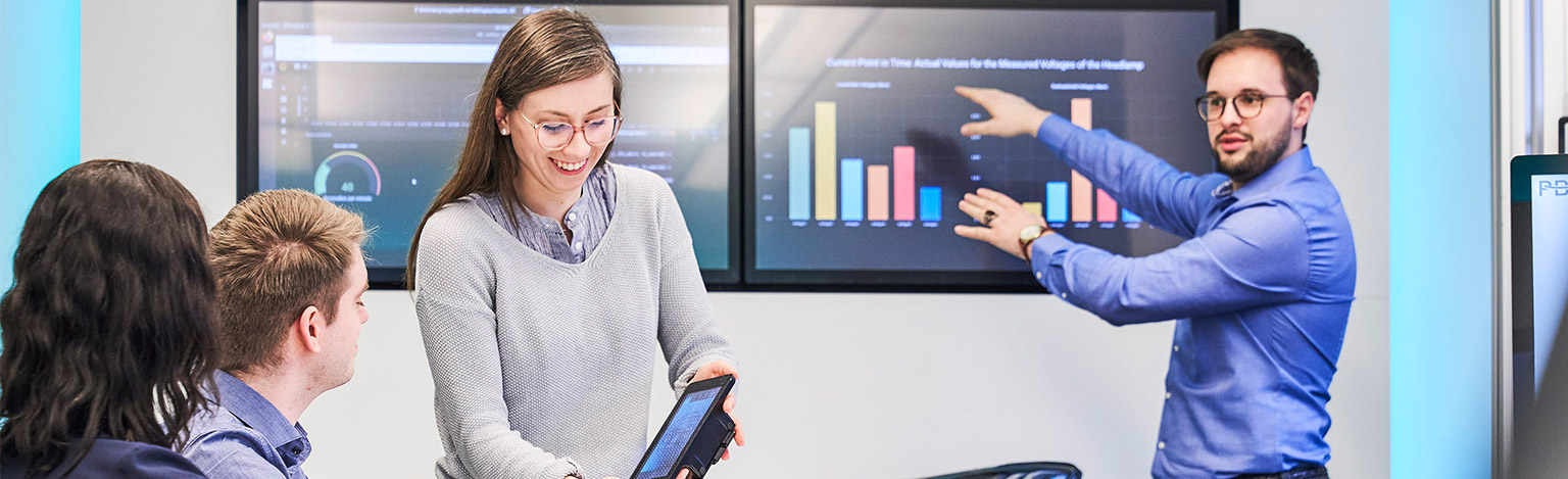 A woman and a man explain a graphic representation displayed on a screen to two other people.