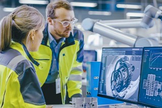 Two people in work clothes at a computer in an industrial hall.